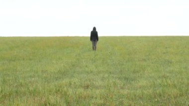 Ufuktaki çayırda haki rengi spor takım elbiseli ve kapüşonlu bir kız duruyor.