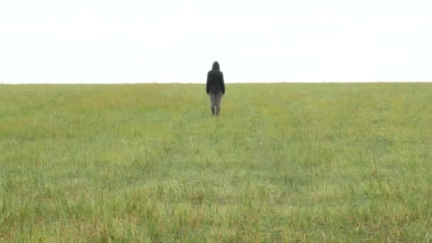 Chica en traje deportivo caqui y capucha se encuentra en un prado en el horizonte — Vídeos de Stock