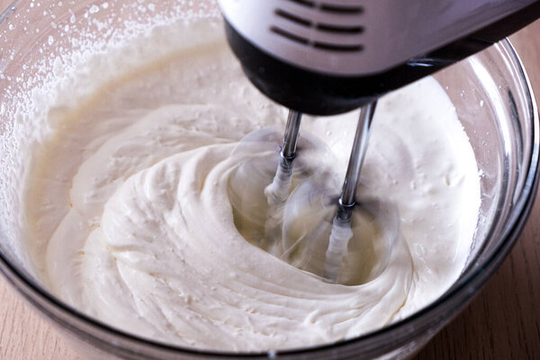 Making whipped cream. Electric hand mixer heating fresh cream in a glass bowl over a wooden surface. 