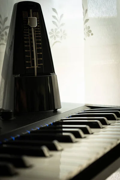 Metronome on top of a piano illuminated by the light coming through the window through the curtain, natural white background, selected focus, music concept — Stock Photo, Image