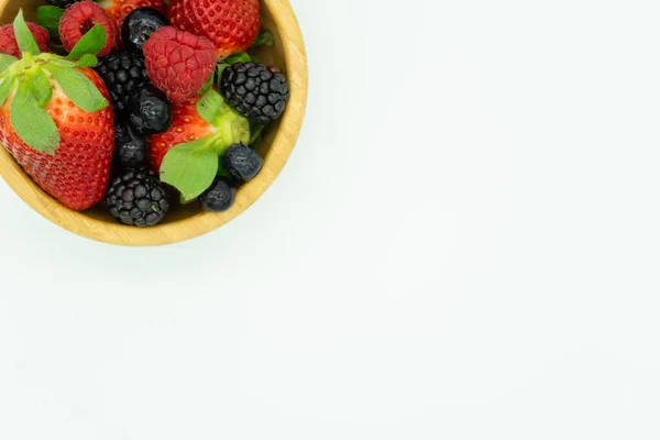 View from above of a wooden bowl filled with strawberries, blackberries, raspberries and blueberries in the upper left corner. White background, space for text. Healthy food concept. — Stock Photo, Image