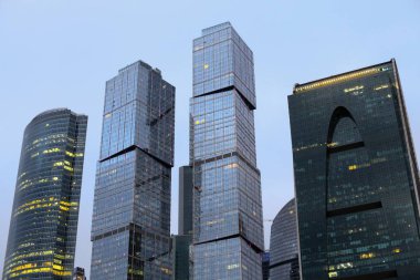Towers of modern high-rise business centers made of glass and concrete against the background of gray sky. Light in windows