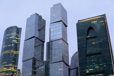 Towers of modern high-rise business centers made of glass and concrete against the background of gray sky. Light in windows