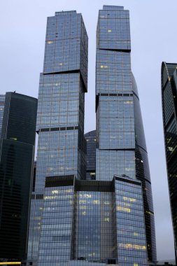 Towers of modern high-rise business centers made of glass and concrete against the background of gray sky. Light in windows