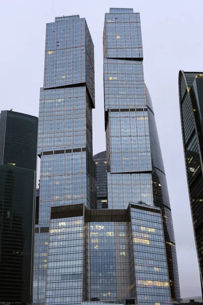 Towers of modern high-rise business centers made of glass and concrete against the background of gray sky. Light in windows
