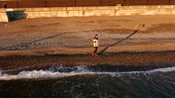 在日落的黄金时刻 年轻的女摄影师带着相机在海滩上散步 美丽的海浪在落日的余晖中冲击着海岸 独立的海上旅行 — 图库视频影像