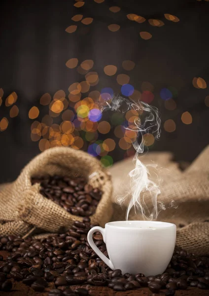 Cangkir kopi dan karung dengan biji kopi di atas meja kayu dan latar belakang bokeh — Stok Foto