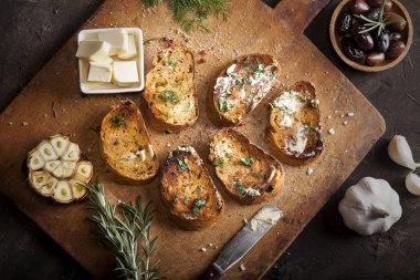 Tereyağlı, zeytinli, sarımsaklı ve otlu dilimlenmiş geleneksel tost ekmeği kesim tahtasında ve karanlık bir masada.