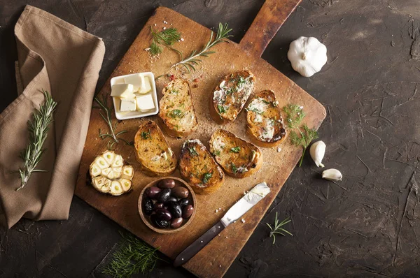 Skivat traditionellt rostat bröd med smör, oliver, vitlök och örter på skärbräda, servett och mörkt bord — Stockfoto