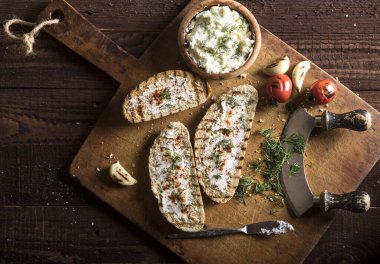 Krem peynirli, zeytinli, sarımsaklı ve kesme tahtasındaki otlu geleneksel tost ekmeği ve karanlık masa. Üst görünüm.