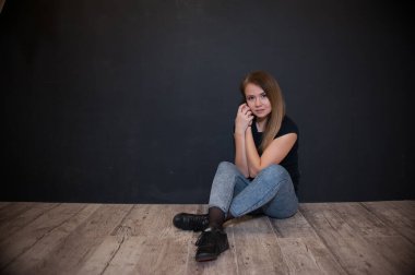 Tişörtlü ve kot pantolonlu bir kız koyu bir arka planda yerde oturuyor. Evde spor yapıyorum.