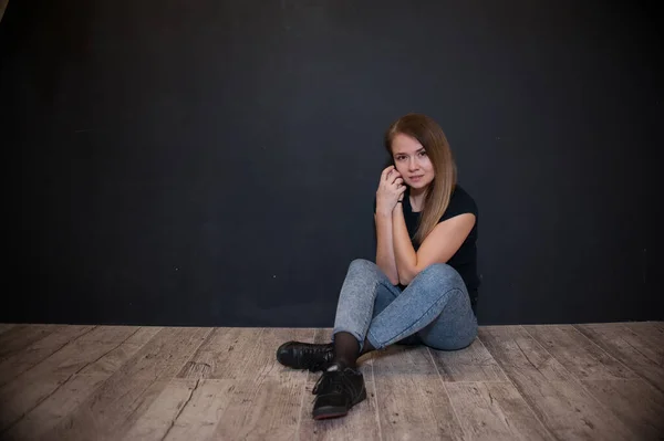 girl in a T-shirt and jeans sits on the floor on a dark background. Doing sports at home