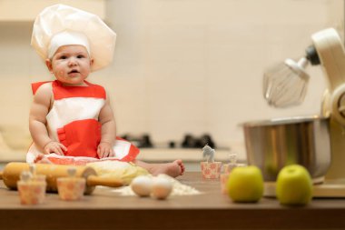 Bir çocuk, bir bebek, bir kız mutfak masasında, bir aşçının şapkasında oturuyor.