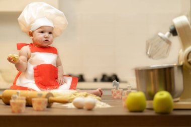 Bir çocuk, bir bebek, bir kız mutfak masasında, bir aşçının şapkasında oturuyor.