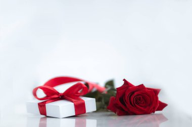 Kırmızı kurdeleli beyaz hediye kutusu ve beyaz arka planda güzel koyu kırmızı bir gül. Noel ve Yeni Yıl tatili kavramı.