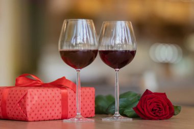 Kırmızı hediye kutusu bir gül ve masada iki kadeh şarap. Kırmızı kurdeleli bir hediye. Sevgililer günü ve kış tatilleri hediye konsepti.