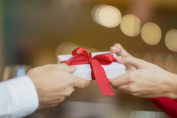 Een man geeft een witte geschenkdoos met een rood lint door aan zijn vriendin. . Warme en mooie achtergrond van een restaurant. Twee glazen wijn en een roos op de tafel van het café. Valentijnsdag concept. — Stockfoto