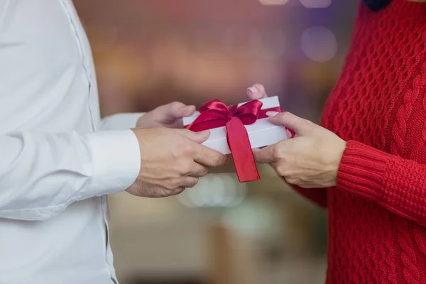 Un hombre pasa una caja de regalos blanca con una cinta roja a las manos de su novia. Aspecto clásico de una pareja, suéter rojo y camisa blanca. Concepto de vacaciones de San Valentín e invierno . — Foto de Stock