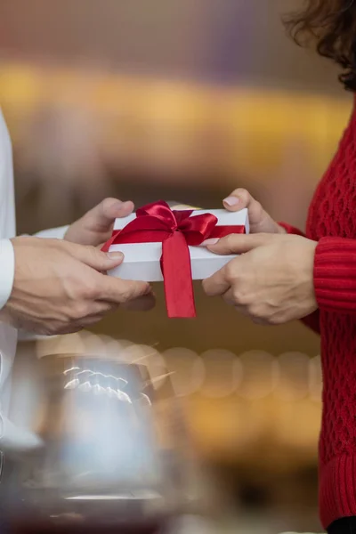 Un uomo passa una scatola regalo bianca con un nastro rosso nelle mani della sua ragazza. Look classico di coppia, maglione rosso e camicia bianca. Giorno di San Valentino e vacanze invernali concetto . — Foto Stock