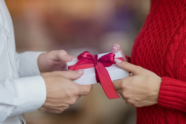 Un uomo passa una scatola regalo bianca con un nastro rosso nelle mani della sua ragazza. Look classico di coppia, maglione rosso e camicia bianca. Giorno di San Valentino e vacanze invernali concetto . — Foto Stock