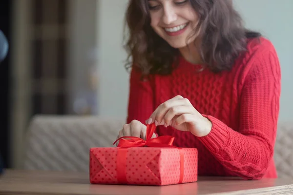Een jonge vrouw opent een cadeautje in een café. Rode geschenkdoos met een lint wordt losgemaakt op de tafel van een restaurant. Valentijnsdag en wintervakantie concept. — Stockfoto