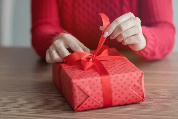 Una joven abre un regalo en un café. Una caja de regalo roja con una cinta está siendo desatada en la mesa de un restaurante. Concepto de vacaciones de San Valentín e invierno . —  Fotos de Stock