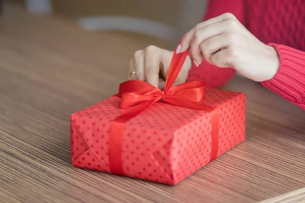 Una joven abre un regalo en un café. Una caja de regalo roja con una cinta está siendo desatada en la mesa de un restaurante. Concepto de vacaciones de San Valentín e invierno . —  Fotos de Stock