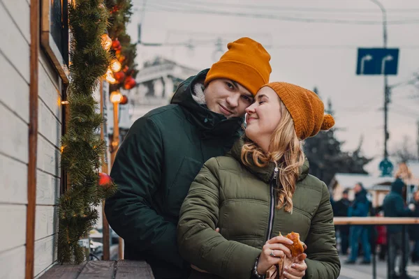 Веселая любящая пара, стоящая на улице, обнимающаяся, целующаяся на размытом праздничном фоне с яркими огнями. Мальчик и девочка в оранжевых шляпах, зеленых куртках. День Святого Валентина и Рождество . — стоковое фото