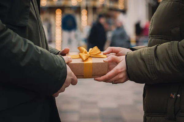 Muž dává malý dárek se žlutou stuhou své přítelkyni na ulici. Žena bere dárek od svého přítele. Valentines day concept. — Stock fotografie