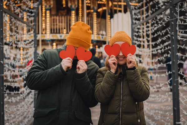 Feliz casal jovem está segurando corações de papel vermelho e sorrindo em um fundo embaçado com luzes laranja na rua de inverno. Conceito de dia dos namorados . — Fotografia de Stock