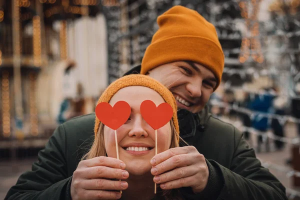 Счастливая молодая пара держит красные бумажные сердечки и улыбается на размытом фоне с оранжевыми огнями на зимней улице. День святого Валентина . — стоковое фото