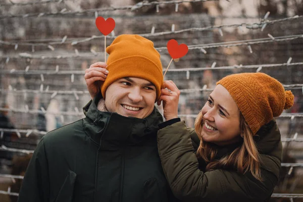 Счастливая молодая пара держит красные бумажные сердечки и улыбается на размытом фоне с оранжевыми огнями на зимней улице. Бумажные черви вонзаются в шляпу. День святого Валентина . — стоковое фото
