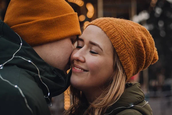 Kochająca piękna para w pomarańczowych kapeluszach i zielonych kurtkach przytula się i całuje na rozmytym tle ze światłami. Zimowe wakacje romantyczne wibracje. Koncepcja Walentynek. — Zdjęcie stockowe