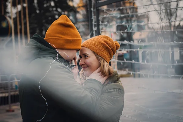 Loving beautiful couple in orange hats and green jackets hug and kiss on the blurry background with lights. Winter holidays romantic vibes. Valentines day concept. — Stock Photo, Image