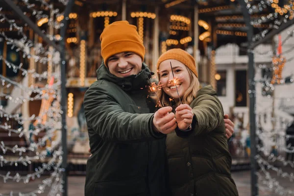 Супруги Красигуль празднуют вместе на рождественском рынке. Девушка и парень с блестками в руках в оранжевых шляпах и зеленых куртках. Концепция зимнего отдыха . — стоковое фото