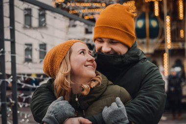 Turuncu şapkalı ve yeşil ceketli genç çift Noel caddesinde şefkatle sarılıp öpüşüyor. Kış tatili konsepti, Sevgililer Günü..