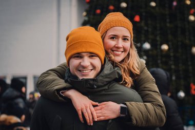 Turuncu şapkalı ve yeşil ceketli genç çift Noel caddesinde şefkatle sarılıp öpüşüyor. Kış tatili konsepti, Sevgililer Günü..