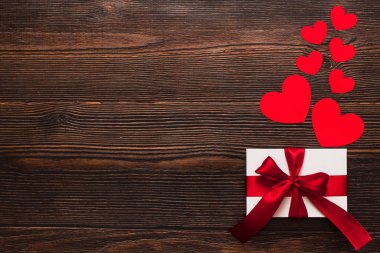 Kırmızı kurdeleli beyaz bir hediye ve koyu ahşap arka planda izole edilmiş kağıt kalpler. Sıcak bir düzleşmeyi kutluyoruz. Sevgililer günü ve Noel konsepti. Kopyalama alanı.