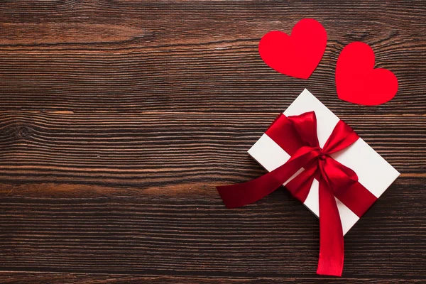 White present with a red ribbon and paper hearts isolated on a dark wooden background. Top side view of a celebrating warm flatlay. Valentines day and Christmas concept. Copyspace. — Stock Photo, Image