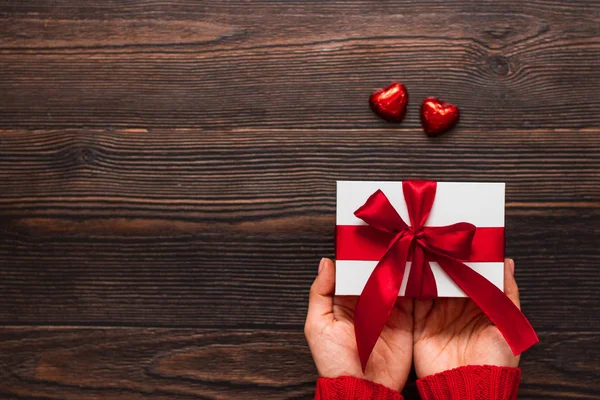 Regalo blanco con una cinta roja en las manos de la mujer y dos dulces de chocolate en forma de corazón sobre un fondo de madera oscura. Concepto de San Valentín. Espacio de copia . —  Fotos de Stock