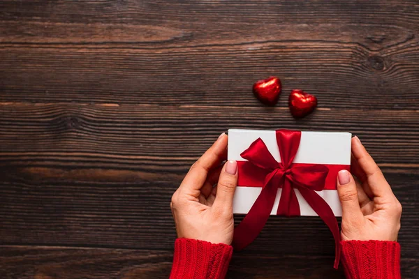Wit cadeau met een rood lint in vrouwenhanden en twee chocolade snoepjes in hartvorm op een donkere houten achtergrond. Valentijnsdag concept. Kopieerruimte. — Stockfoto