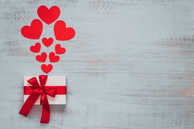Paper hearts and a craft present with red ribbon on a light painted wooden background. Top side angle view, flat lay. Valentines day concept. Copyspace.