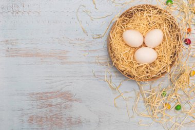 Üç beyaz yumurta ve bir kutuda saman yuvasında renkli yuvarlak şekerler. Açık mavi ahşap zemin üzerinde düz bir üst görünüm. İfade için kopyalama uzayı. Paskalya tatili konsepti.