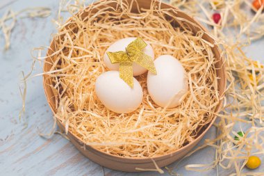 Altın yaylı üç beyaz yumurta ve saman yuvasında renkli yuvarlak şekerler. Açık mavi ahşap arka planda üst taraf manzarası. İfade için kopyalama uzayı. Paskalya tatili konsepti.