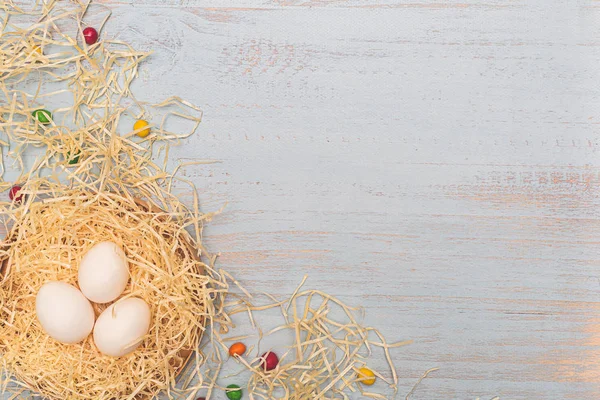 Três ovos brancos e doces redondos coloridos em um ninho de feno em uma caixa. Vista lateral superior plana sobre um fundo de madeira azul claro. Copyspace para a redação. Conceito feriados Páscoa . — Fotografia de Stock