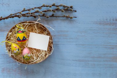 Saman yuvasında renkli paskalya yumurtaları ve içinde mavi ahşap arka planda izole edilmiş söğüt ağacından yapılmış boş bir kart. Paskalya tatili konsepti. Kopyalama alanı, metin ve sözcükler için yer. Nisan titreşimleri