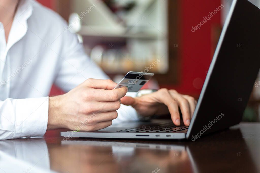 Closeup of hands of young businessman wearing a white fashion shirt with a credit card and laptop making payment online in a cafe. Freelance and selfemployment concept. Distance job.