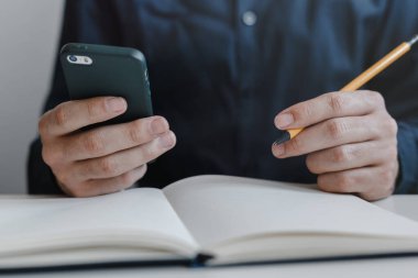 Adamın elleri cep telefonu ve parmaklarının arasında bir kalem tutarken. Defter masanın üstünde. Ofiste çalışan genç bir iş adamı. İş, finans, endüstri ve iş konsepti