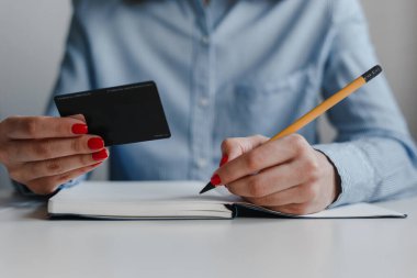 Not defterine kırmızı tırnaklı, sarı kalemli ve elinde mavi gömlekli bir kadın eli. İş, serbest meslek, serbest meslek. Uzaklık işi ve çevrimiçi iş kavramı