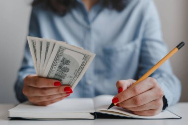 Kırmızı tırnaklı, elinde 100 dolarlık banknotlar olan ve mavi gömlekli bir kalemle not alan kadınların yakınına. İş, serbest çalışma. Uzaklık işi ve çevrimiçi iş kavramı